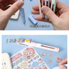 A pink folding pen-shaped safety scissor is shown resting on a sheet of craft paper with various stickers and drawings.