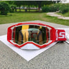 A pair of red and orange motocross goggles resting on a white paper on a gray surface.