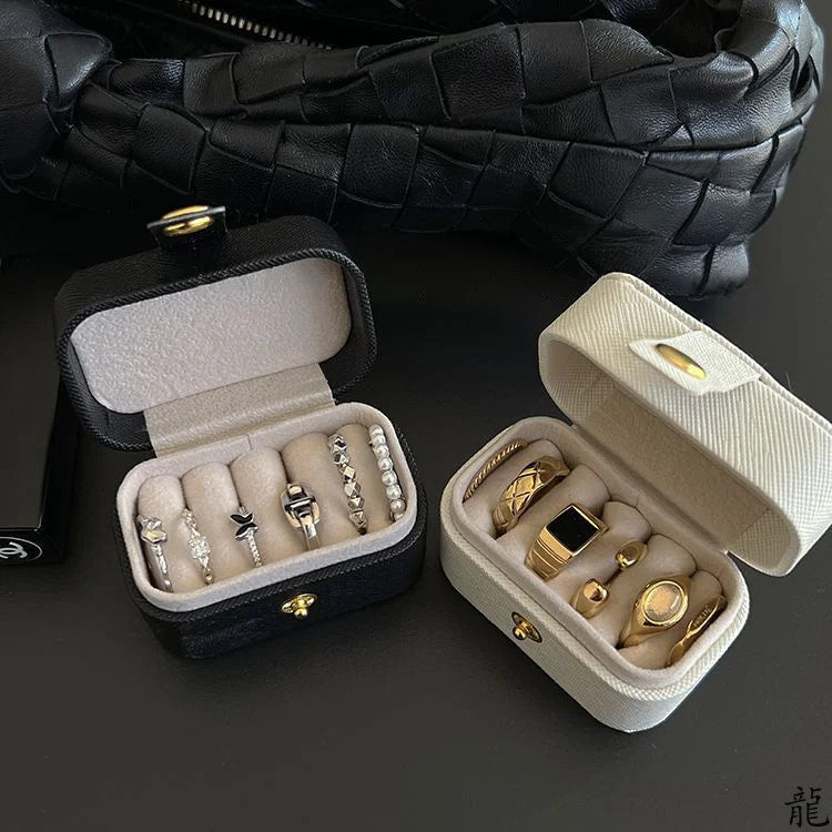 A black leather jewelry travel case is shown open, revealing several gold and silver rings and a bracelet inside.