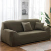 A modern, taupe elastic stretch sofa cover is displayed indoors next to a window.