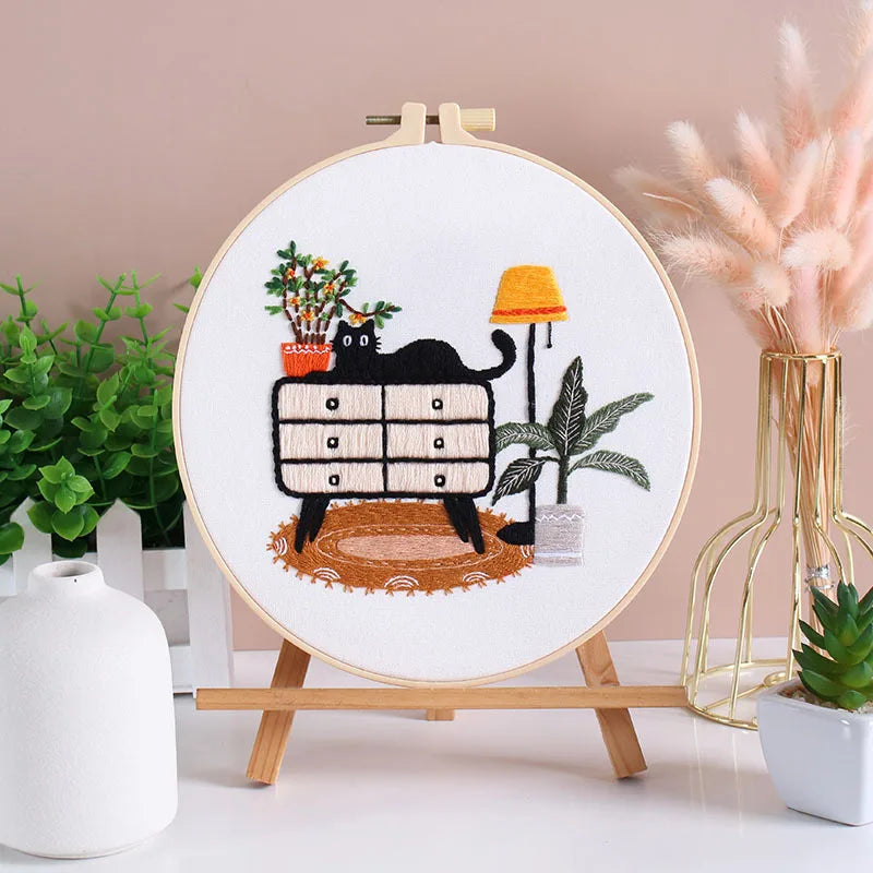 A circular embroidery hoop displaying a black cat and floral designs on white fabric, set against a light pink background.