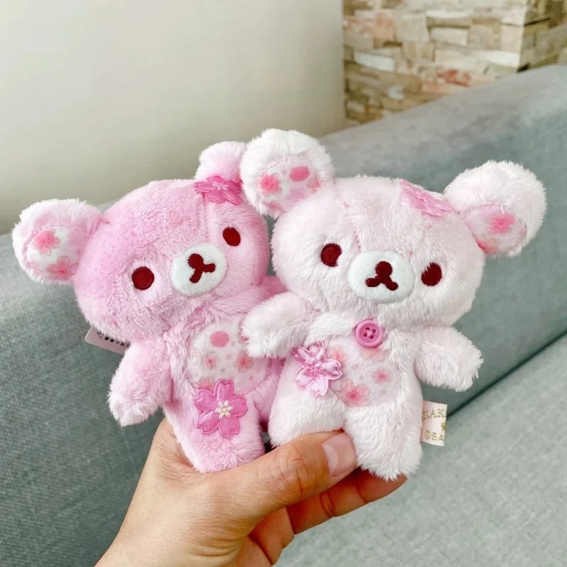 A pink cherry blossom plush bear keychain is held in a hand against a gray sofa background.