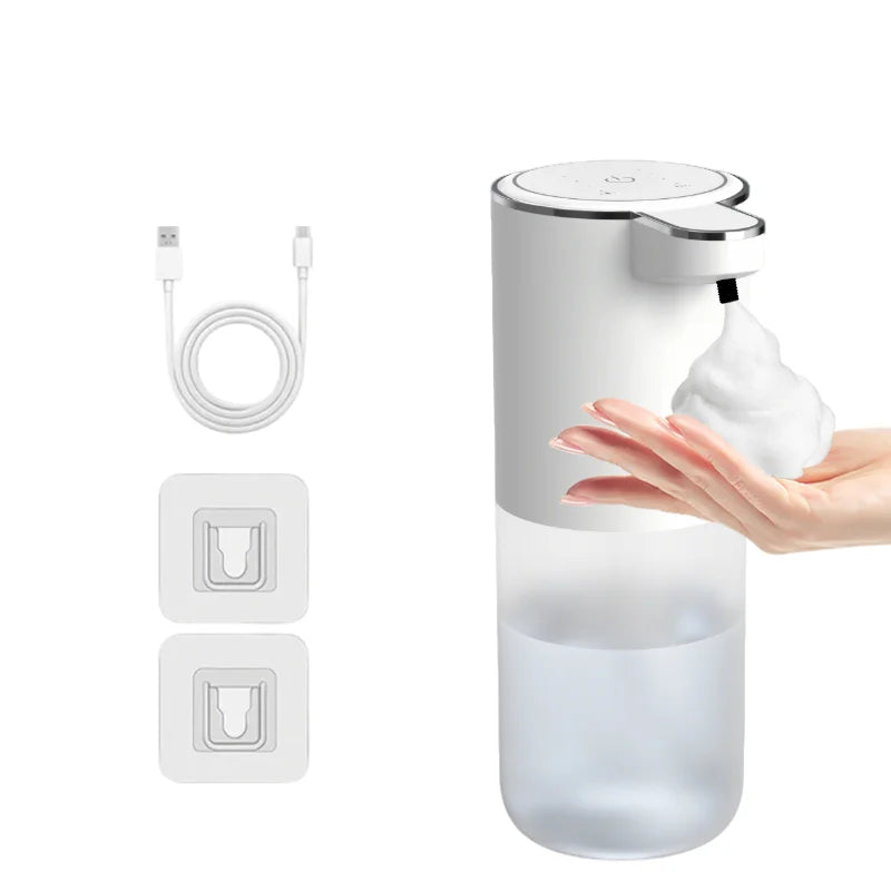A white touchless automatic soap dispenser with a pump head and a clear liquid reservoir, shown next to its charging cable an