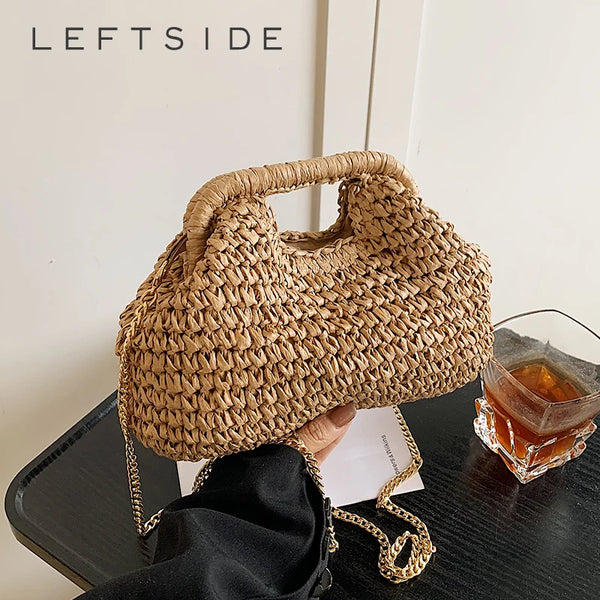A woven straw chain crossbody handbag is held against a dark surface next to an amber glass.
