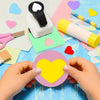 Hands holding a yellow heart-shaped paper punch tool on a blue crafting surface surrounded by colorful paper scraps.
