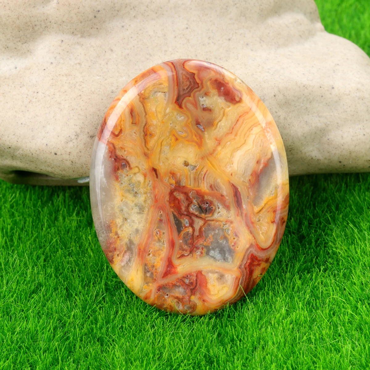 A polished, oval natural crystal stone with swirling shades of orange, red, and yellow rests on green grass.