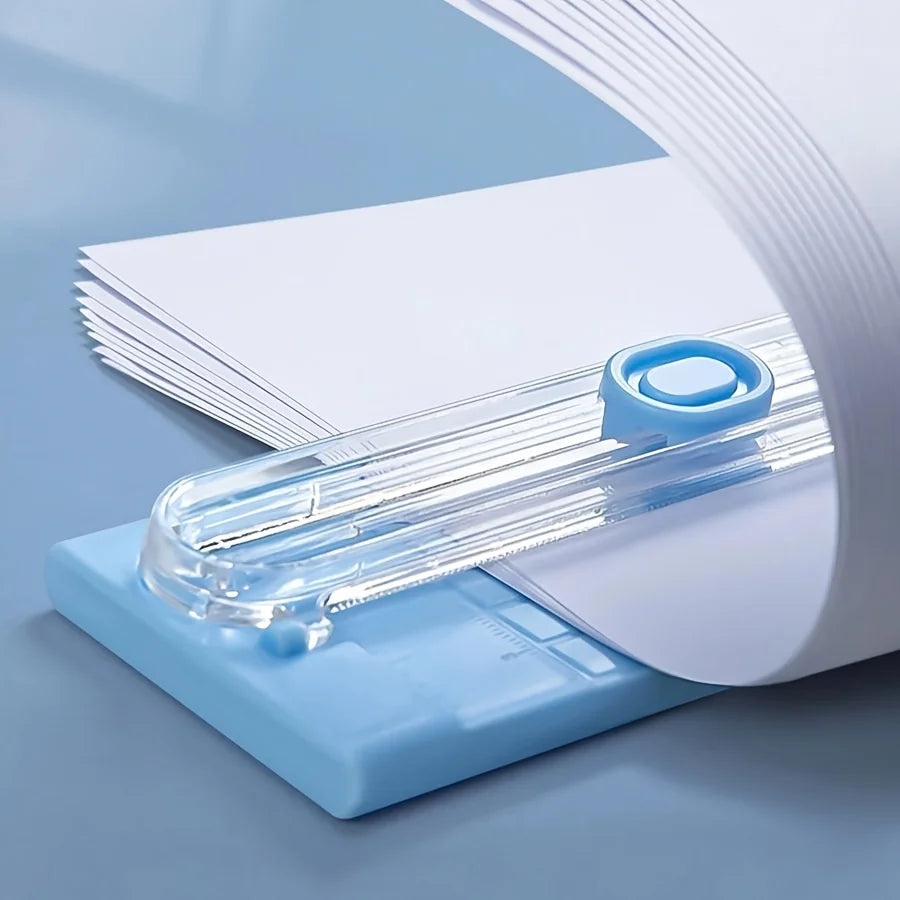 A close-up view of a portable paper cutter trimmer with a clear plastic blade and blue casing, positioned over a stack of whi