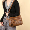 A woman wearing a black sweater and gray blazer holds a brown leather tote bag with an adjustable strap.