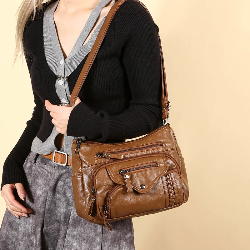 A woman wearing a black sweater and gray blazer holds a brown leather tote bag with an adjustable strap.