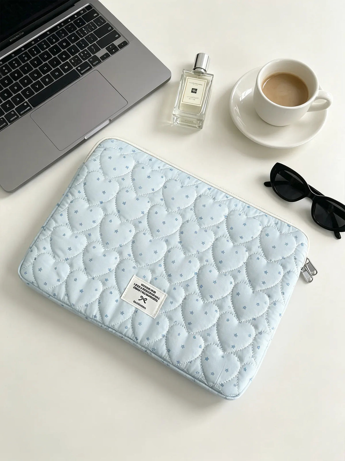 A light blue quilted laptop sleeve with a soft lining resting on a white surface next to a cup and sunglasses.