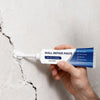 A hand holds a tube of white wall repair paste, labeled 8.8 oz e 250g, against a cracked white wall.