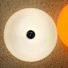 A white, spherical LED wall sconce with a black center is shown against a woven background and an orange light source.