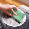 A hand is using a green double-sided kitchen scouring sponge on a metal dish.