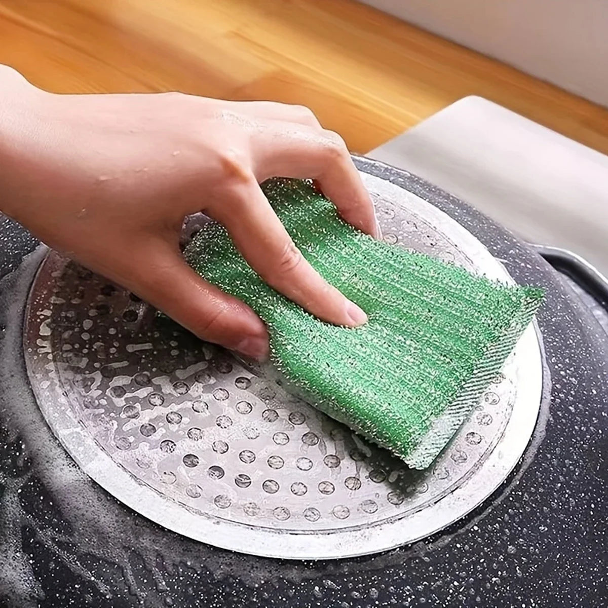 A hand is using a green double-sided kitchen scouring sponge on a metal dish.