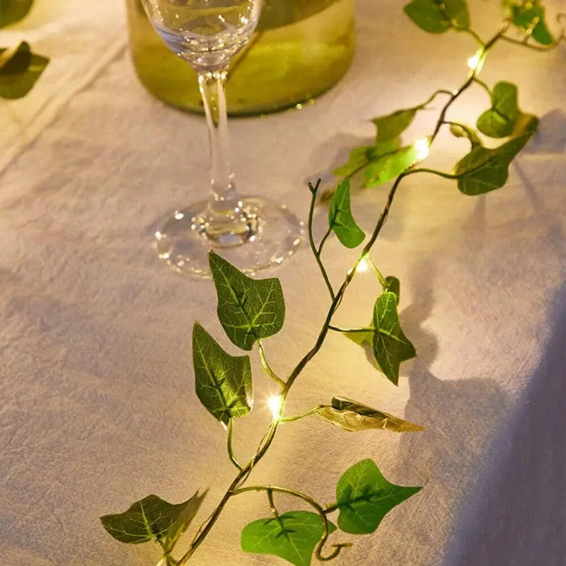Une guirlande d'ivy vert avec des lumières LED chaudes traîne sur un tissu blanc à côté d'un verre.