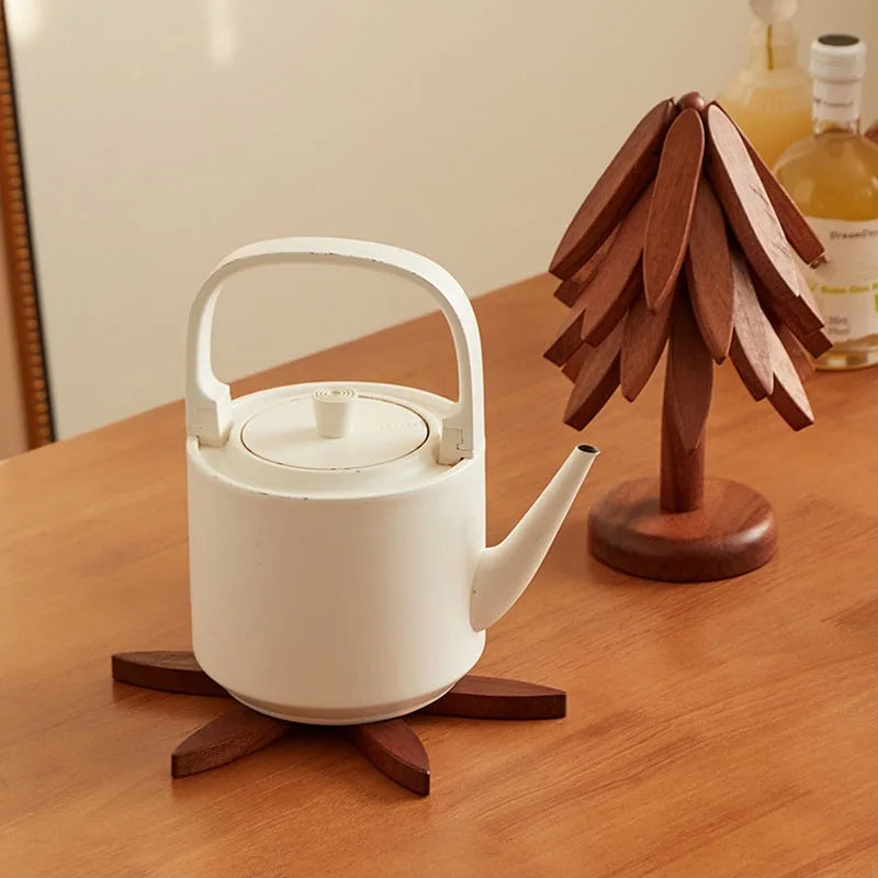 A white ceramic teapot sits on a wooden trivet next to a wooden tree-shaped object and a bottle.