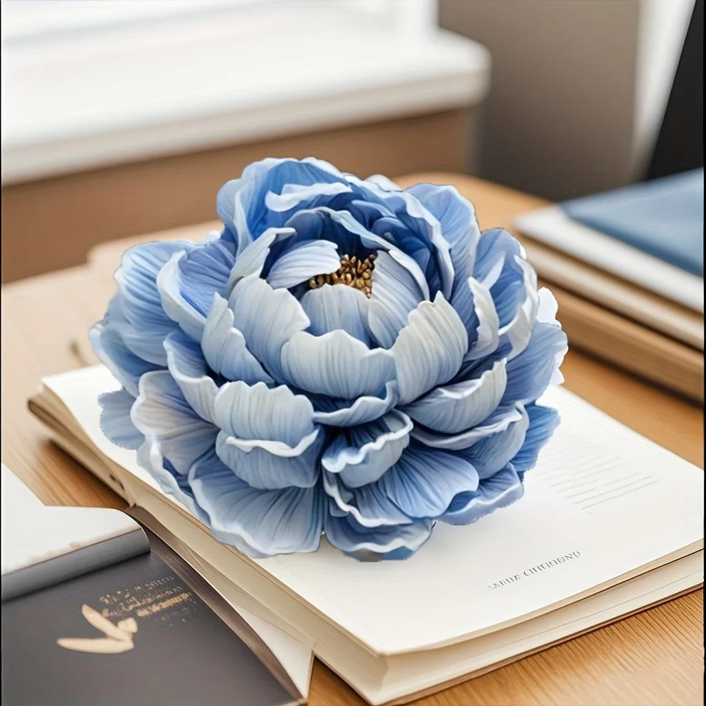 A light blue, fully bloomed peony flower made from flexible silicone mold resting on an open notebook.
