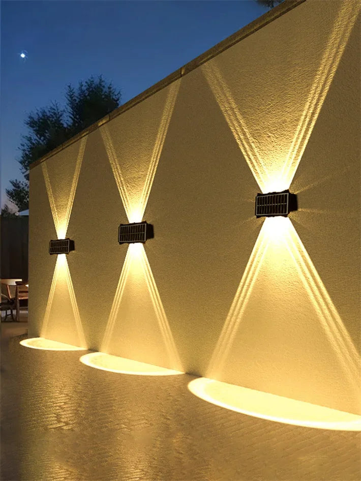 A solar powered LED wall light illuminates a textured wall with upward and downward beams at dusk.