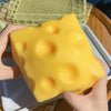 A hand holds a large, yellow, squishy cheese fidget toy with dimples against a white plate.