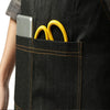 A black denim work apron pocket holds a silver smartphone and two yellow metal rings.