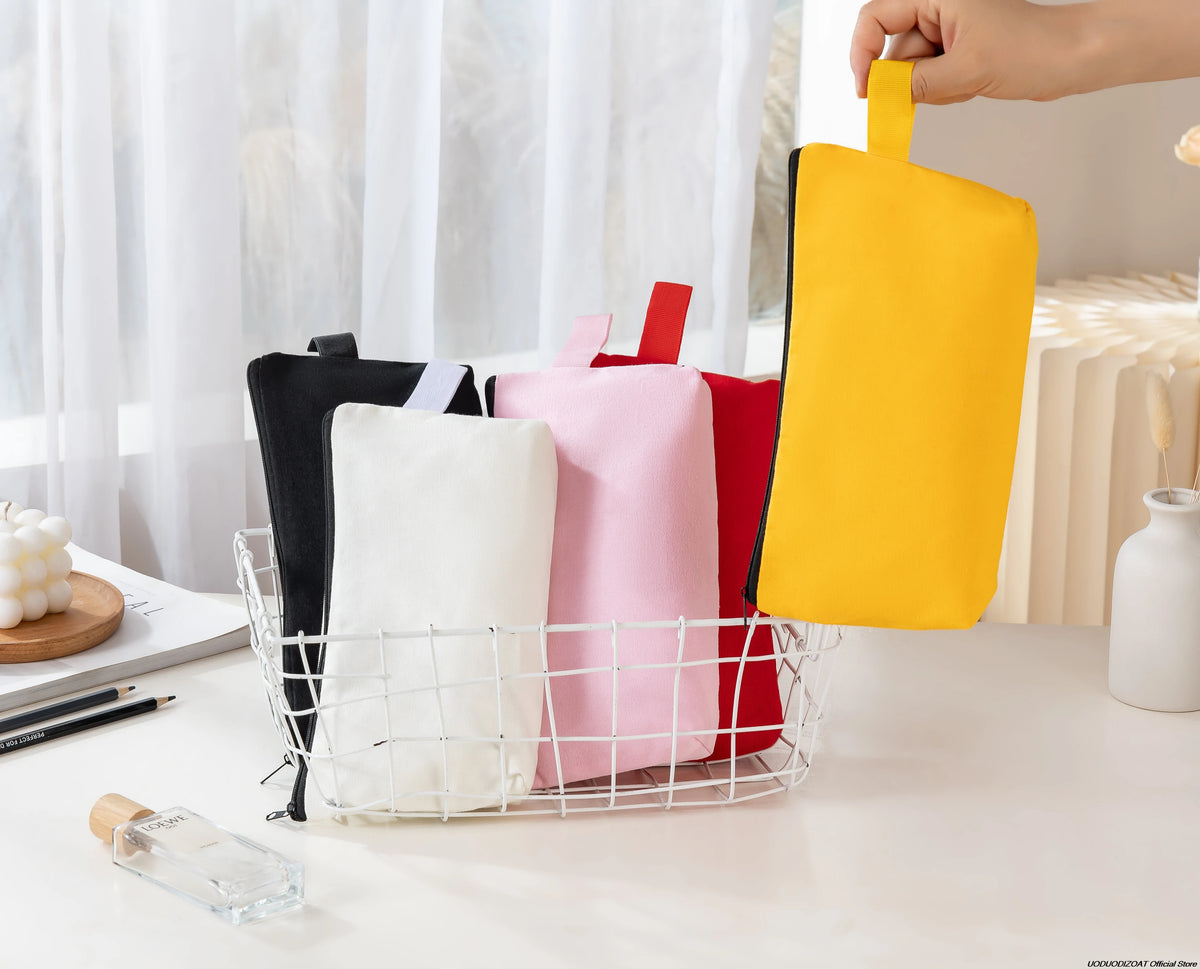 A hand is holding a bright yellow canvas zipper pouch next to several pink and red pouches in a white wire basket.