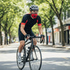 A man wearing a black cycling jersey and helmet rides a bicycle on a city street while wearing a red face mask.