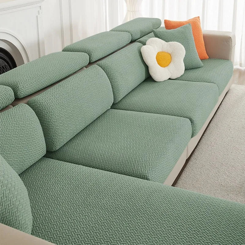 A light sage green jacquard elastic sofa slipcover with orange accent pillows on a white sofa.