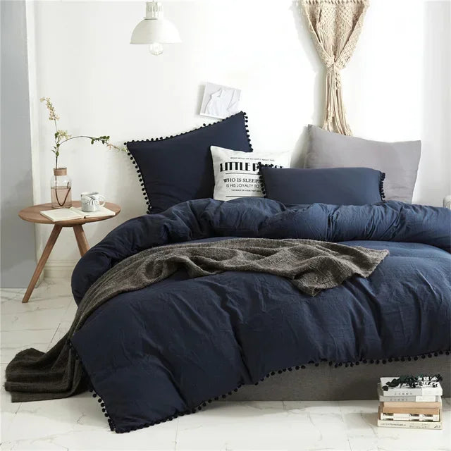 A dark blue duvet cover set with pom tassels and a gray throw blanket on a bed.