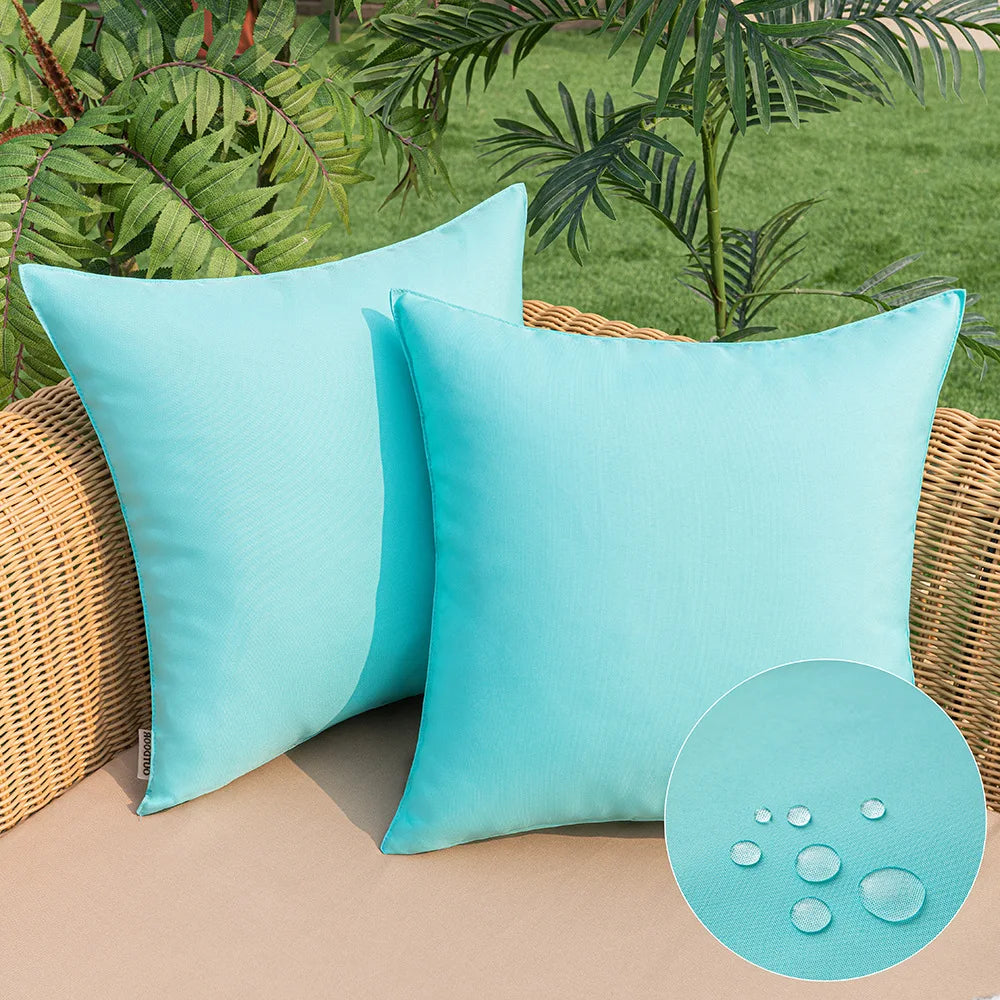 Two light blue linen-textured outdoor pillow covers resting on a tan surface with green foliage in the background.