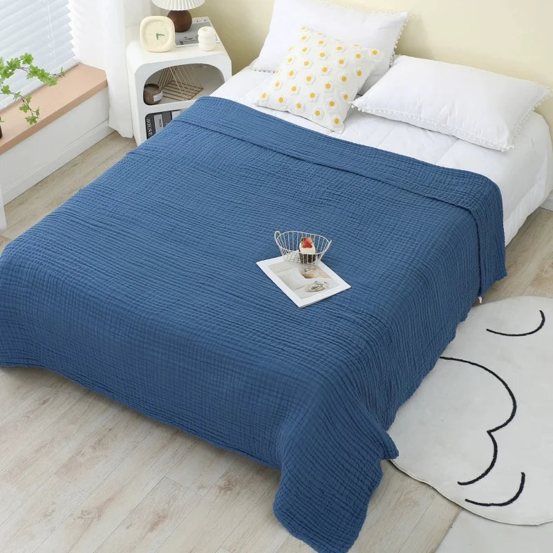 A deep blue pure cotton muslin throw blanket draped over a white bed with yellow patterned pillows.