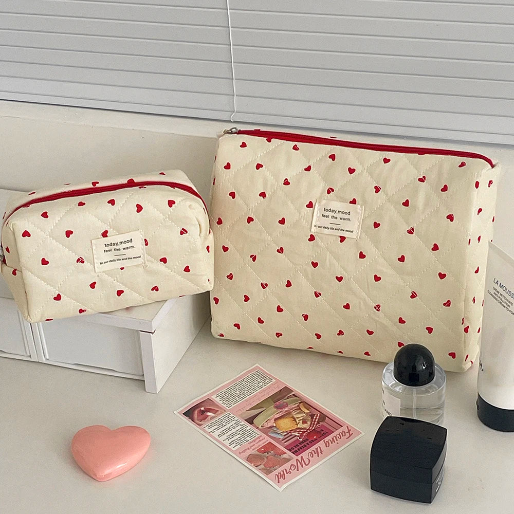 A cream-colored quilted makeup bag with a red heart pattern is displayed next to a pink heart and a small black accessory.