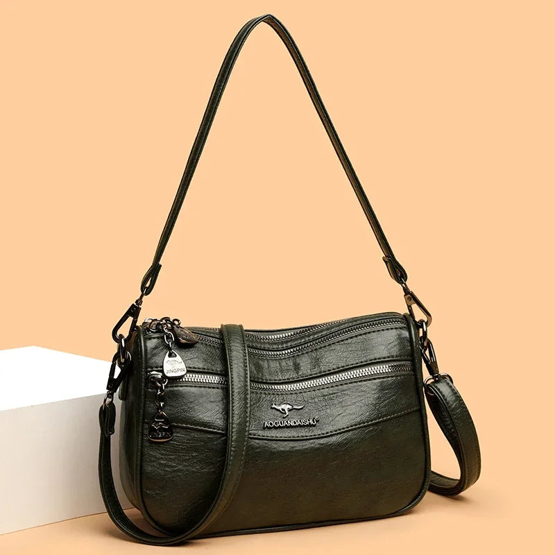 A dark green leather compact square crossbody bag with a long strap resting on a light surface.