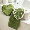 A green leaf-shaped coral fleece hand towel with a white trim and hanging loop resting on a white bathroom sink.
