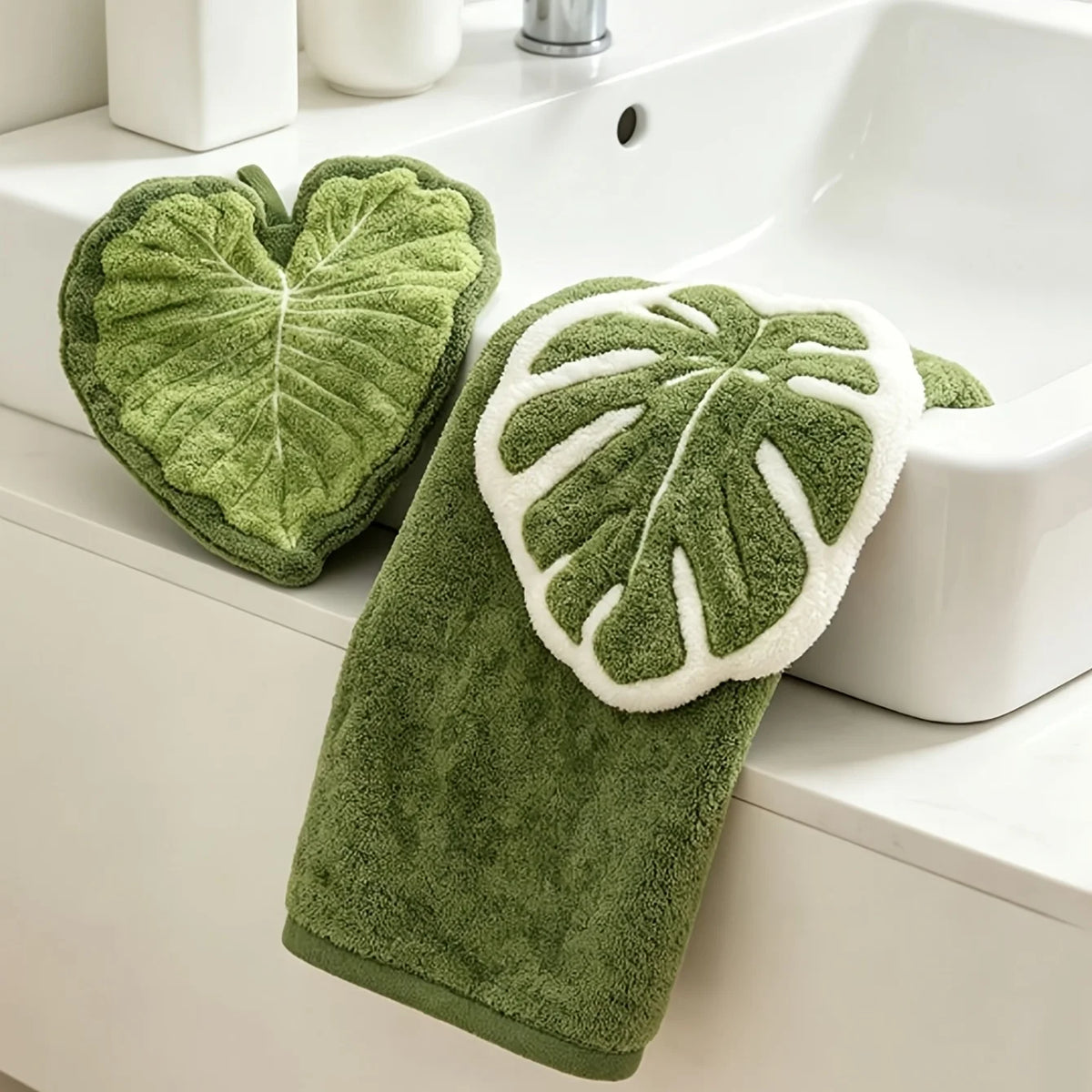 A green leaf-shaped coral fleece hand towel with a white trim and hanging loop resting on a white bathroom sink.