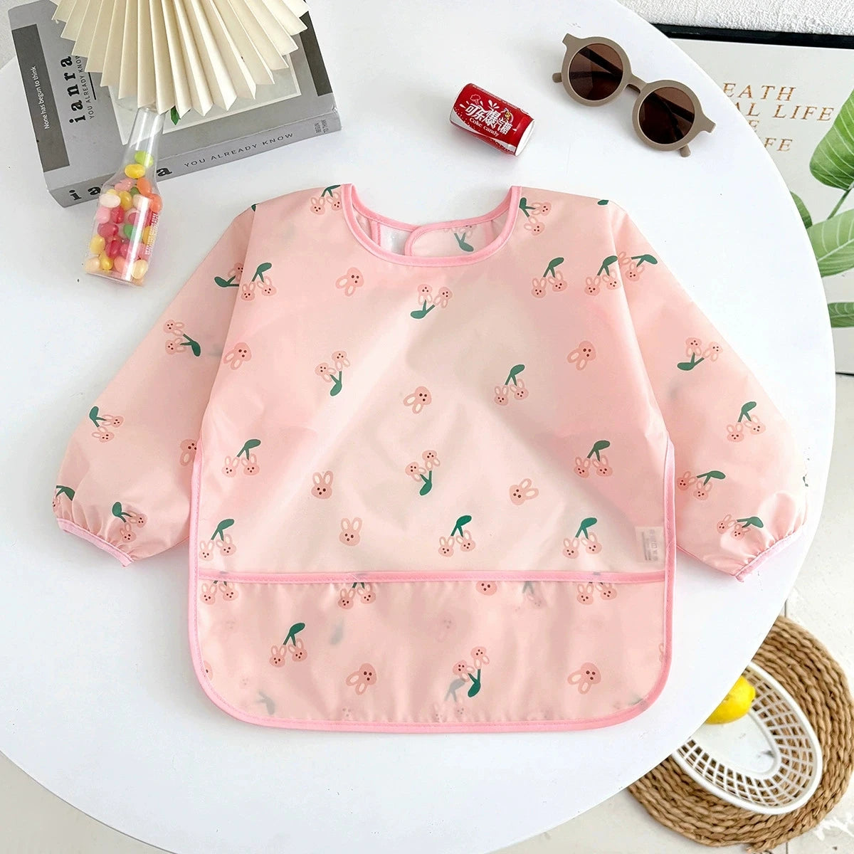A pink waterproof long-sleeve baby feeding smock with a floral pattern displayed on a white table.