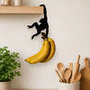 A black monkey silhouette is hanging from a wooden wall hook, which is holding a bunch of yellow bananas.