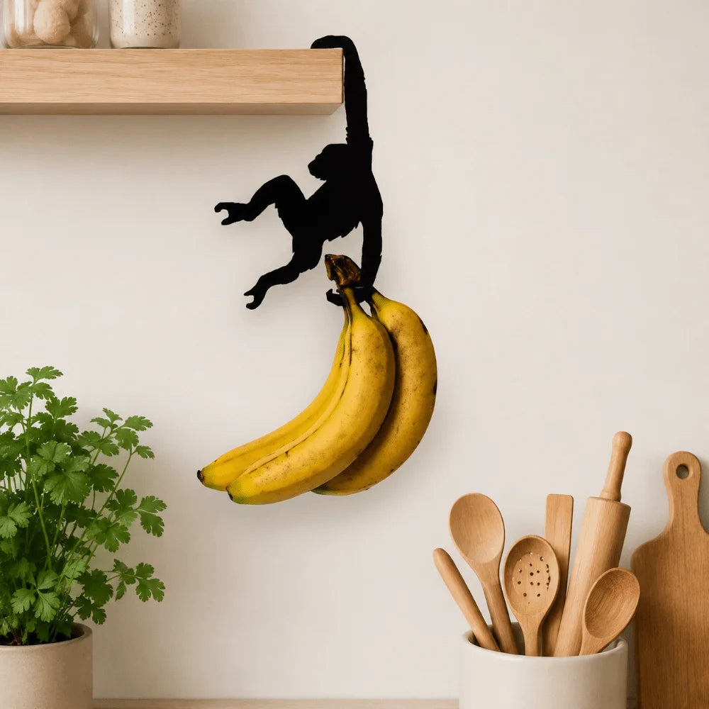 A black monkey silhouette is hanging from a wooden wall hook, which is holding a bunch of yellow bananas.
