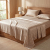 A beige striped satin bed sheet set is draped over a wooden bed frame in a bright bedroom setting.