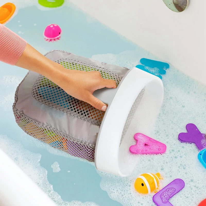 A hand is placing a white hanging bath toy storage bag with a mesh net into a tub filled with soapy water and colorful bath t