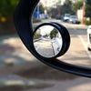 A close-up view of a black dual-lens car blind spot mirror reflecting a street and a white vehicle.