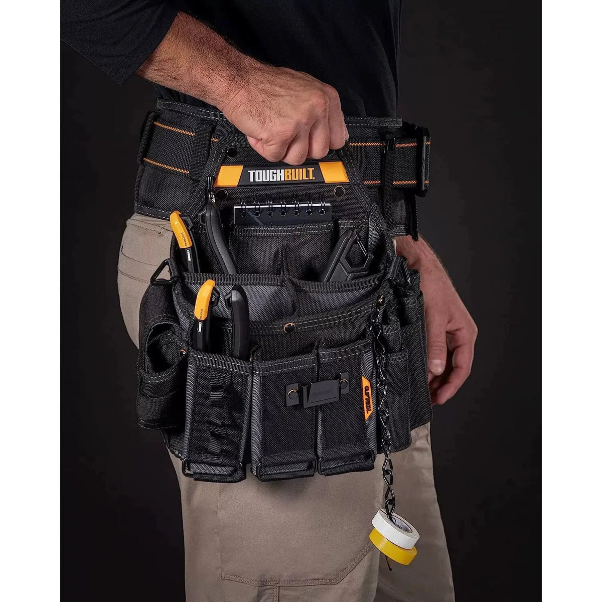 A hand is holding a black ToughBuilt tool belt with multiple pockets against a dark background.