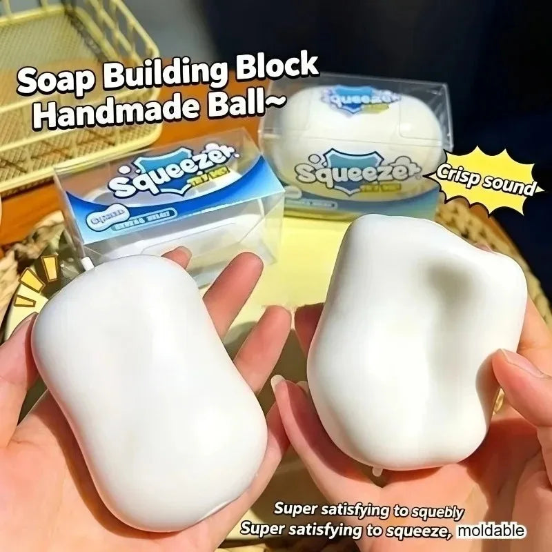 A hand holds a white squishy building block ball next to its packaging and a squeeze toy.