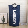 A navy blue foldable laundry bag with a padded handle hanging against a white brick wall.