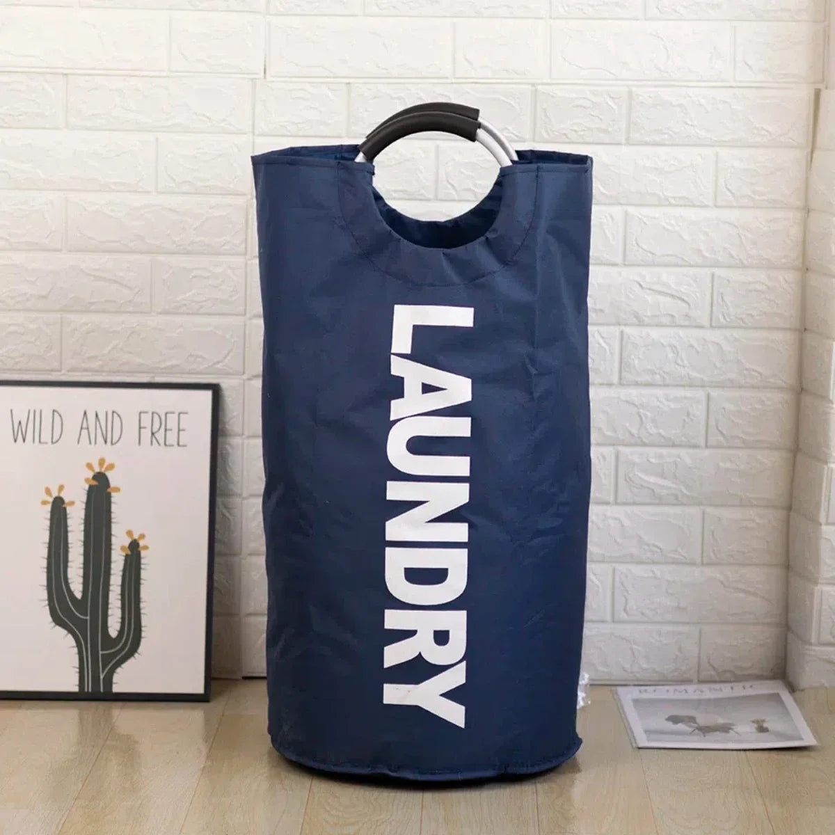 A navy blue foldable laundry bag with a padded handle hanging against a white brick wall.