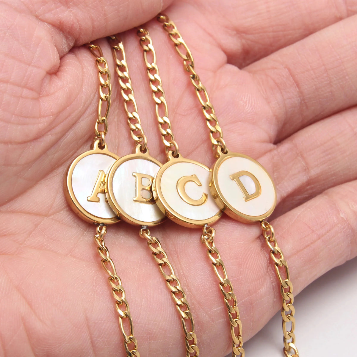 A set of gold-toned stainless steel initial letter bracelets, including A, B, C, and D, displayed on a hand.