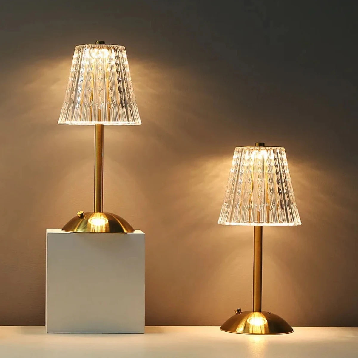 A modern gold-toned table lamp with a clear textured glass shade illuminates a white surface.
