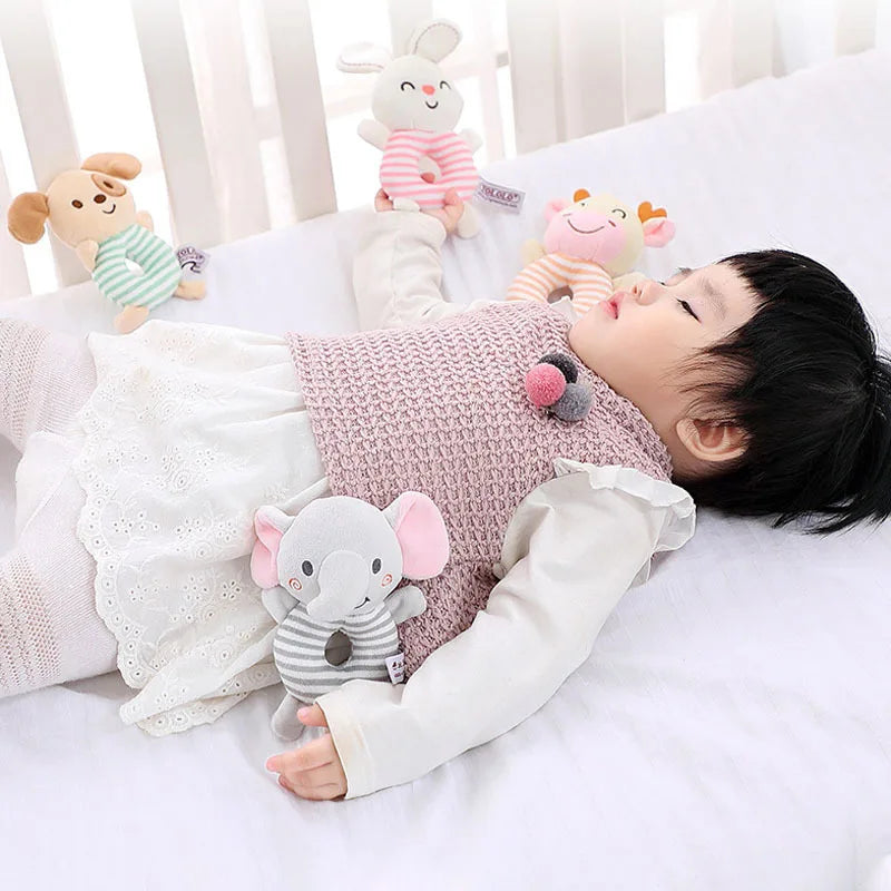 A child is lying on a white bed with several soft plush baby rattle toys featuring cartoon animals like a bear, elephant, and