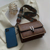 A brown leather mini cross-body bag with a patterned strap and a gold glass ornament, placed on a cream fabric.