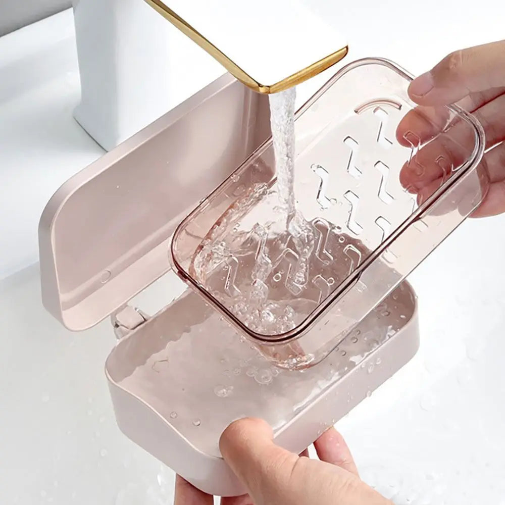A hand is pouring water into a transparent soap dish with a drainage tray, which is part of a pink bathroom accessory.