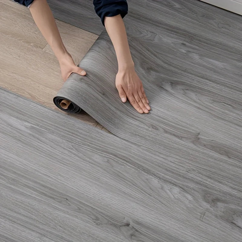 Hands are unrolling a roll of gray wood grain wallpaper onto a floor.